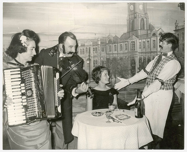 Elizabeth Connelly, TWA air hostess from New York serenaded by head waiter with Mr & Mrs George Miller playing the violin and accordion respectively, Music Hall Restaurant, Apr 1965
